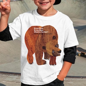 May include: A white t-shirt with a brown bear illustration from the children's book "Brown Bear, Brown Bear, What Do You See?" by Bill Martin Jr. and Eric Carle. The shirt has black sleeves and is worn by a child.