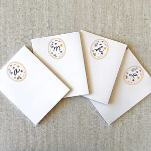 May include: Four white cards with floral and initial designs. Each card features a different initial inside a gold oval, surrounded by colorful flowers. The cards are arranged on a neutral linen surface.