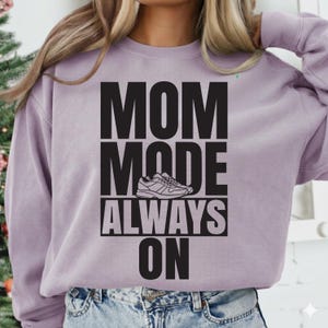 May include: A lilac-colored sweatshirt with the text "MOM MODE ALWAYS ON" in bold black letters. A graphic of a sneaker is between "MODE" and "ALWAYS". The sweatshirt is a casual style.