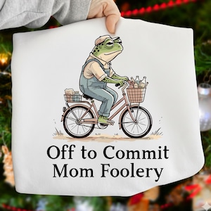 May include: White t-shirt with a cartoon frog riding a bicycle, wearing overalls and a cap. The bicycle has a basket filled with bottles. Text on the shirt reads "Off to Commit Mom Foolery."