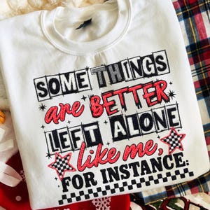 Puede incluir: Sudadera blanca con la frase bordada "SOME THINGS are BETTER LEFT ALONE like me, FOR INSTANCE." El texto es en negro, rojo y gris, con estrellas a cuadros. La sudadera tiene cuello redondo.