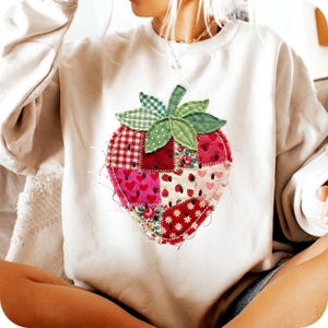Può includere: Felpa color crema con un grande design a patchwork a forma di fragola. La fragola è composta da vari modelli di tessuto, tra cui quadretti rossi, stampe floreali e motivi a forma di cuore. La fragola ha foglie verdi e un gambo.
