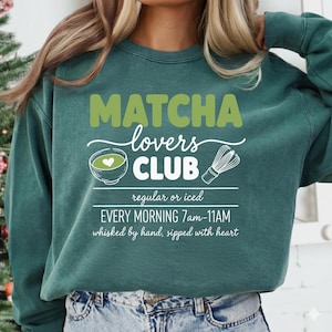May include: Teal green sweatshirt with the text "MATCHA Lovers CLUB" in green and white. The design includes a matcha bowl with a heart, a whisk, and the text "regular or iced" and "EVERY MORNING 7am-11AM".
