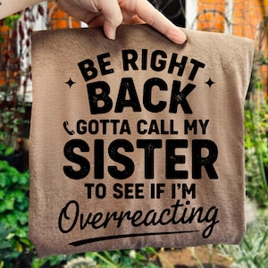 May include: A brown t-shirt with the text "BE RIGHT BACK GOTTA CALL MY SISTER TO SEE IF I'M Overreacting" in black. The text is centered and in a bold, easy-to-read font. The shirt is held up by a hand.