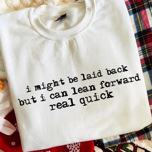 Op de afbeelding: Witte sweatshirt met de zwarte tekst "i might be laid back but i can lean forward real quick". De sweatshirt heeft een ronde hals en lange mouwen. De achtergrond bevat een ruitpatroon en andere decoratieve elementen.