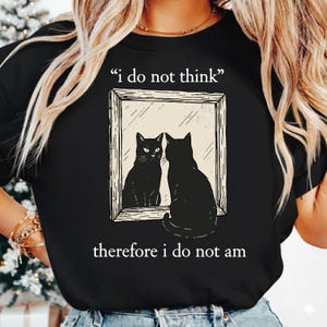 May include: Black t-shirt featuring a graphic of a black cat looking in a mirror at its reflection. The mirror image and the cat are in a rectangular frame. The text above the image reads "i do not think" and the text below reads "therefore i do not am."