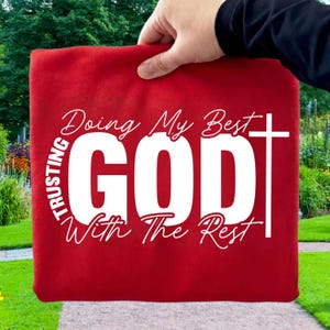 Op de afbeelding: Een rood textiel item met de woorden "TRUSTING GOD" in een cirkelvormig ontwerp en "Doing My Best With The Rest" in witte letters. Een wit kruis staat aan de rechterkant van de tekst. Het item wordt door een hand omhoog gehouden.
