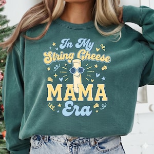 Könnte beinhalten: Ein teal-grünes Sweatshirt mit dem Text "In My String Cheese Mama Era" in einem Retro-Schriftzug. Die Grafik zeigt einen Käse-Stick mit Sonnenbrille, umgeben von Sternen und Käseformen.