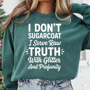 May include: A teal green sweatshirt with the text "I DON'T SUGARCOAT I Serve Raw TRUTH With Glitter And Profanity" in white. The sweatshirt is a solid color and has a crew neck.