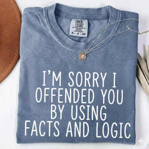 Op de afbeelding: Een blauw Comfort Colors t-shirt met de witte tekst "I'M SORRY I OFFENDED YOU BY USING FACTS AND LOGIC". Een zilveren ketting met een hanger ligt over het shirt.