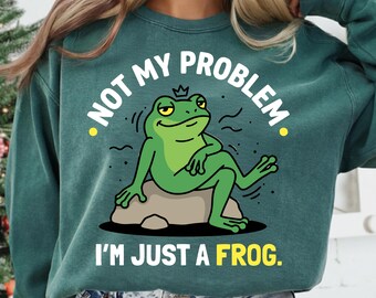 May include: Teal green sweatshirt with the text "NOT MY PROBLEM" and "I'M JUST A FROG." A cartoon frog wearing a crown is lounging on a rock. The frog is green with yellow eyes and a smug expression.