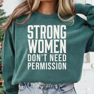 Op de afbeelding: Een teal groene sweatshirt met de witte tekst "STRONG WOMEN DON'T NEED PERMISSION". De sweatshirt heeft een ronde hals en lange mouwen. De afbeelding is een close-up van het bovenlichaam.