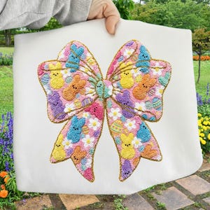 May include: A white fabric item featuring a large, embroidered bow design. The bow is filled with colorful Easter-themed elements, including Peeps, Easter eggs, and flowers. The bow is outlined in gold thread.
