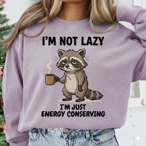 May include: A light purple sweatshirt with the text "I'M NOT LAZY I'M JUST ENERGY CONSERVING". The graphic features a cartoon raccoon holding a coffee mug, with steam rising from it. The raccoon has a tired expression.