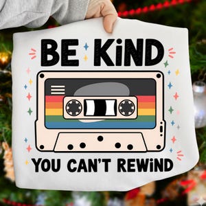 May include: White graphic sweatshirt with a retro cassette tape design. The cassette tape features a rainbow stripe and the words "BE KIND YOU CAN'T REWIND" in black lettering. The design is surrounded by colorful star accents.