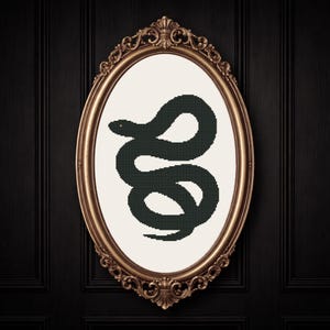 May include: An oval framed cross-stitch art piece featuring a dark green snake with a red eye on a white background. The frame is gold-colored with ornate detailing, set against a black paneled wall.