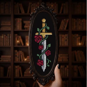 May include: Oval framed cross-stitch art featuring a sword entwined with roses and vines. The sword has a gold hilt and a silver blade. The roses are red, and the vines are green. The background is black.