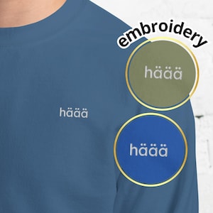 Peut inclure: Un sweat-shirt bleu avec le mot "häää" brodé en blanc sur la poitrine. Le mot "broderie" est écrit en arc de cercle au-dessus de deux écussons circulaires, un vert et un bleu, chacun avec le mot "häää".