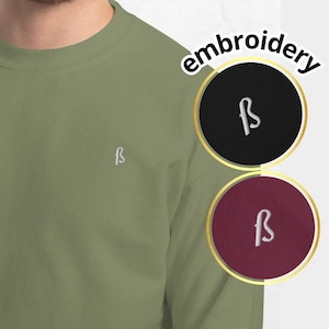 May include: An olive green sweatshirt featuring a small white embroidered "f b" logo. The image also displays two embroidery options: a black circle with a white "f b" logo and a burgundy circle with a white "f b" logo.