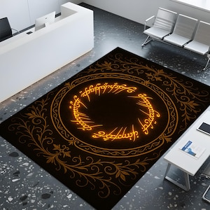 May include: A rectangular black rug featuring a golden ring design with script. The rug has a dark brown center and is surrounded by a golden border with floral accents. The rug is placed on a gray floor.