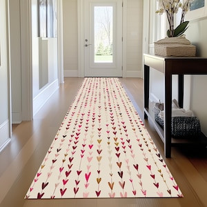 May include: A long, rectangular runner rug with a cream background and a repeating pattern of vertical lines of hearts in shades of pink, red, and brown. The rug is placed in a hallway with a wooden floor.