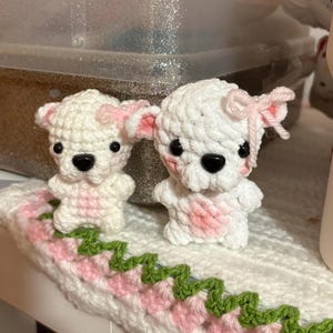 May include: Two small, white crocheted animal figures with pink accents on their ears and chests. Each figure features black plastic eyes and a black nose, with a light pink yarn bow attached to one ear. They are positioned on a white crocheted base.