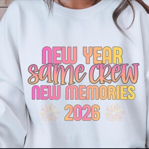 Puede incluir: Sudadera blanca con el texto "NEW YEAR SAME CREW NEW MEMORIES 2026" en rosa, naranja y amarillo. El diseño incluye gráficos de fuegos artificiales. Una prenda casual y festiva.