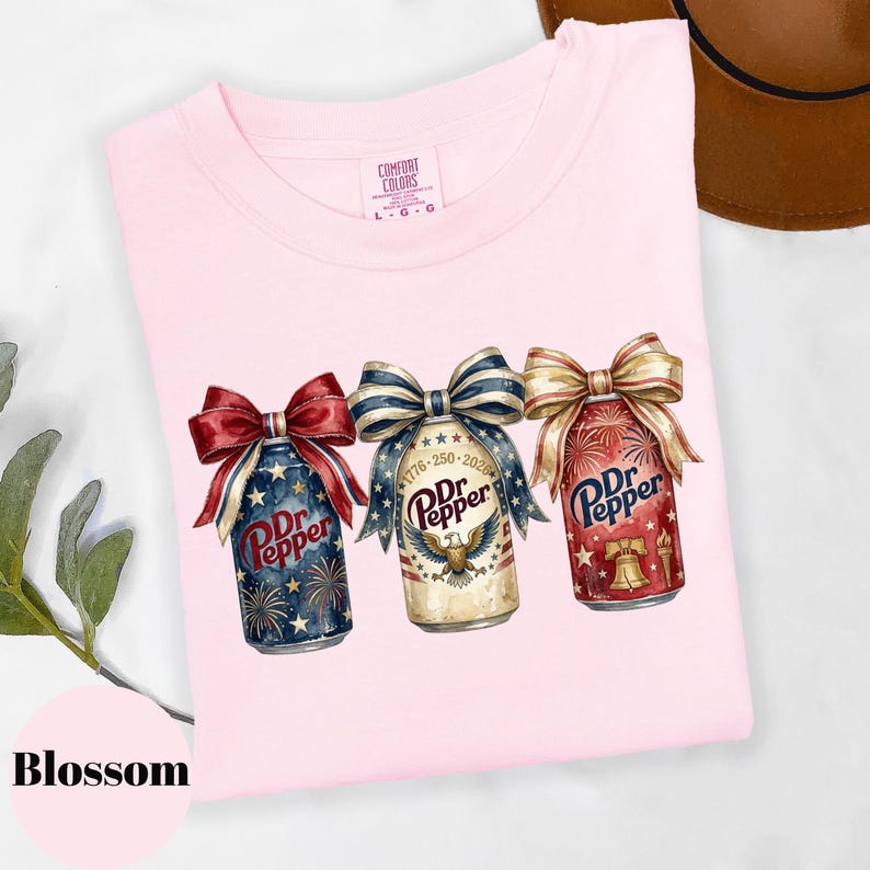 Puede incluir: Camiseta rosa claro Comfort Colors con tres latas de Dr. Pepper adornadas con lazos patri&oacute;ticos. Las latas muestran fuegos artificiales, estrellas y el texto "1776-250-2026". La palabra "Blossom" est&aacute; en la parte inferior izquierda.