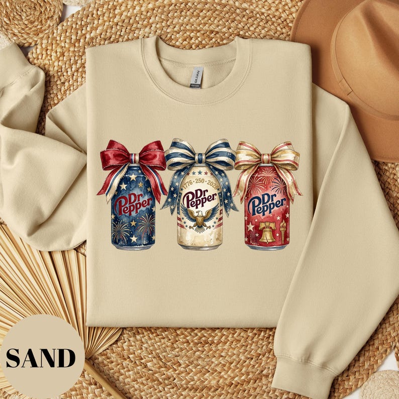 Puede incluir: Sudadera color arena con tres latas de Dr. Pepper con dise&ntilde;os de lazos patri&oacute;ticos. Las latas est&aacute;n decoradas con fuegos artificiales, un &aacute;guila y una campana de la libertad. El texto "Dr. Pepper" es visible en cada lata. La palabra "SAND" est&aacute; impresa en la parte inferior izquierda.