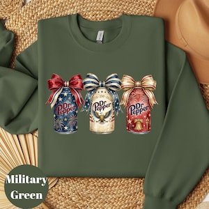 Puede incluir: Sudadera verde militar con tres dise&ntilde;os de latas de Dr. Pepper, cada una adornada con lazos patri&oacute;ticos. Las latas muestran colores rojo, blanco y azul, fuegos artificiales y el texto "Dr. Pepper". Un estilo de sudadera c&oacute;modo e informal.