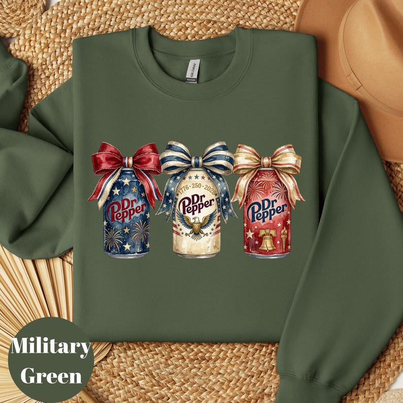 Puede incluir: Sudadera verde militar con tres dise&ntilde;os de latas de Dr. Pepper, cada una adornada con lazos patri&oacute;ticos. Las latas muestran colores rojo, blanco y azul, fuegos artificiales y el texto "Dr. Pepper". Un estilo de sudadera c&oacute;modo e informal.