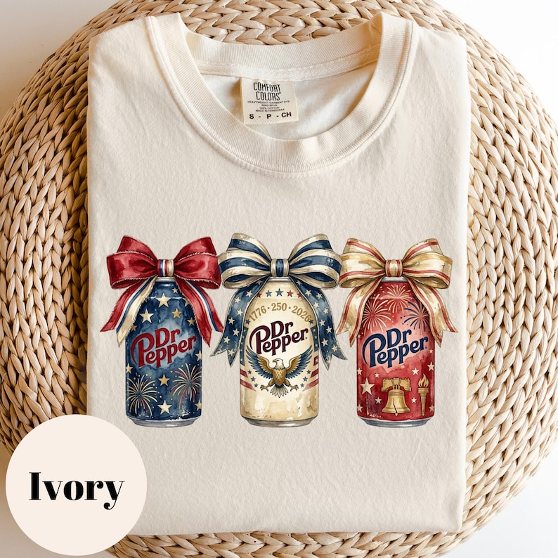 Puede incluir: Camiseta color marfil con tres dise&ntilde;os de latas de Dr. Pepper adornadas con lazos patri&oacute;ticos. Las latas muestran fuegos artificiales, un &aacute;guila y la Campana de la Libertad. La camiseta tiene cuello redondo y mangas cortas.