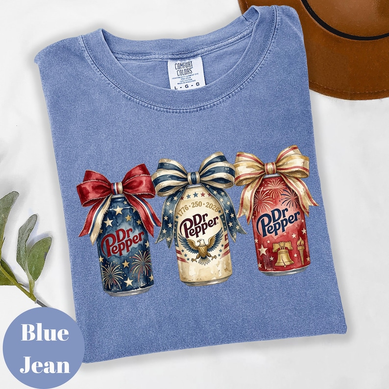Puede incluir: Camiseta azul vaquero con tres latas de Dr. Pepper adornadas con lazos patri&oacute;ticos. Las latas presentan dise&ntilde;os de fuegos artificiales y estrellas. Una lata tiene el texto "1776-250-2026". La camiseta tiene una etiqueta Comfort Colors.