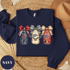 Puede incluir: Sudadera azul marino con tres dise&ntilde;os de latas de Dr. Pepper, cada uno con un lazo patri&oacute;tico. Las latas muestran fuegos artificiales, un &aacute;guila y una campana de la libertad. La sudadera tiene la palabra "NAVY" en un c&iacute;rculo azul marino.