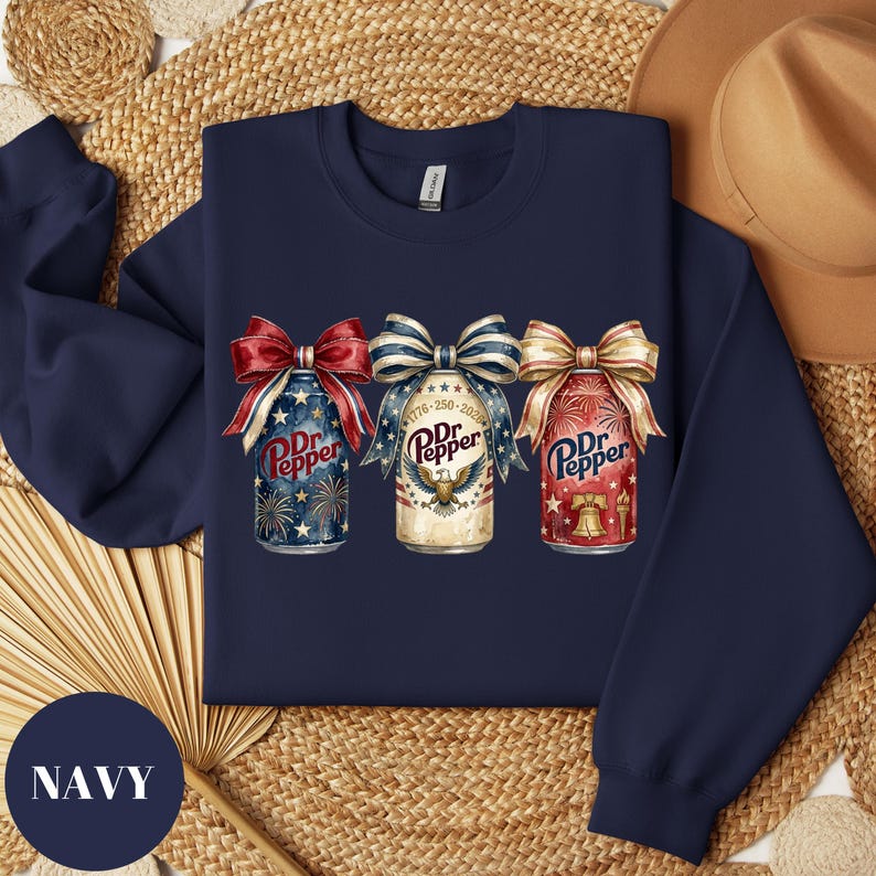 Puede incluir: Sudadera azul marino con tres dise&ntilde;os de latas de Dr. Pepper, cada uno con un lazo patri&oacute;tico. Las latas muestran fuegos artificiales, un &aacute;guila y una campana de la libertad. La sudadera tiene la palabra "NAVY" en un c&iacute;rculo azul marino.