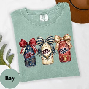 Puede incluir: Camiseta verde claro con tres latas de Dr. Pepper decoradas con lazos patri&oacute;ticos. Las latas presentan dise&ntilde;os de fuegos artificiales, un &aacute;guila y una campana, con el texto "Dr. Pepper".