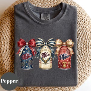 Puede incluir: Camiseta gris oscuro con tres dise&ntilde;os de latas de Dr. Pepper con lazos patri&oacute;ticos. Las latas est&aacute;n decoradas con fuegos artificiales, un &aacute;guila y la Campana de la Libertad. La camiseta est&aacute; hecha de un material suave y c&oacute;modo.