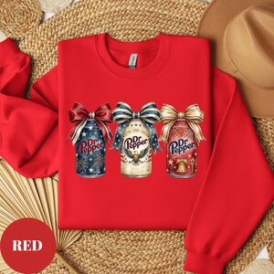 Puede incluir: Sudadera roja con tres dise&ntilde;os de latas de refresco Dr. Pepper con lazos patri&oacute;ticos. Las latas tienen fuegos artificiales, un &aacute;guila y un gr&aacute;fico de campana. El texto "Dr. Pepper" es visible. La palabra "RED" est&aacute; en un c&iacute;rculo rojo.