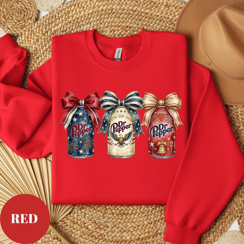 Puede incluir: Sudadera roja con tres dise&ntilde;os de latas de refresco Dr. Pepper con lazos patri&oacute;ticos. Las latas tienen fuegos artificiales, un &aacute;guila y un gr&aacute;fico de campana. El texto "Dr. Pepper" es visible. La palabra "RED" est&aacute; en un c&iacute;rculo rojo.