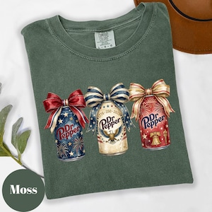 Puede incluir: Camiseta verde musgo Comfort Colors con tres dise&ntilde;os de latas de Dr. Pepper con lazos patri&oacute;ticos. Las latas est&aacute;n decoradas con fuegos artificiales, estrellas y el texto "Dr. Pepper".
