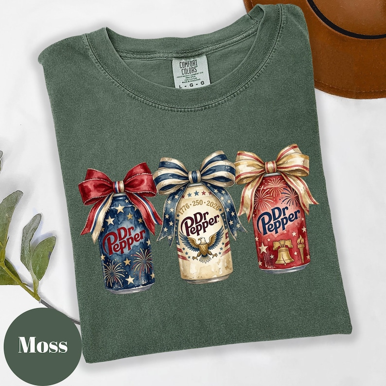 Puede incluir: Camiseta verde musgo Comfort Colors con tres dise&ntilde;os de latas de Dr. Pepper con lazos patri&oacute;ticos. Las latas est&aacute;n decoradas con fuegos artificiales, estrellas y el texto "Dr. Pepper".