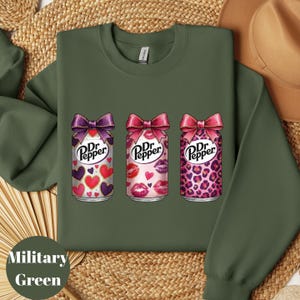 May include: Military green sweatshirt featuring three Dr. Pepper cans with bows. The cans have different designs: hearts, lips, and leopard print. The text "Dr. Pepper" is visible on each can.