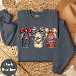 Puede incluir: Sudadera gris oscuro con tres dise&ntilde;os de latas de Dr. Pepper, cada una adornada con un lazo patri&oacute;tico. Las latas muestran fuegos artificiales, estrellas y los colores rojo, blanco y azul. El texto "Dr. Pepper" es visible.