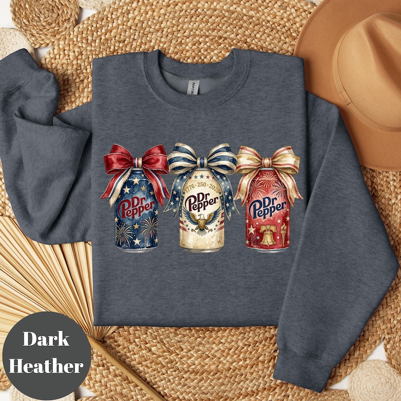Puede incluir: Sudadera gris oscuro con tres dise&ntilde;os de latas de Dr. Pepper, cada una adornada con un lazo patri&oacute;tico. Las latas muestran fuegos artificiales, estrellas y los colores rojo, blanco y azul. El texto "Dr. Pepper" es visible.