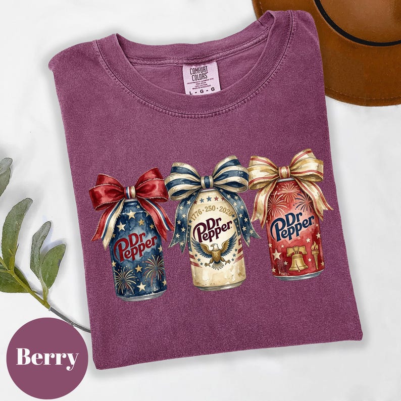 Puede incluir: Camiseta color bayas con tres latas de Dr. Pepper con dise&ntilde;os de lazos patri&oacute;ticos. Las latas tienen ilustraciones de fuegos artificiales y campanas. La camiseta tiene una etiqueta Comfort Colors.