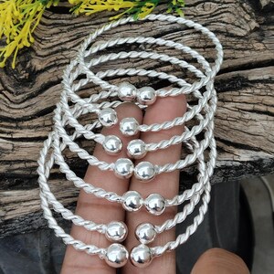 Könnte beinhalten: Eine Sammlung silberfarbener Armbänder. Jedes Armband hat ein gedrehtes Seildesign und eine zentrale silberne Perle. Die Armbänder werden auf einer Holzoberfläche präsentiert, mit ein paar Zweigen gelber Blumen im Hintergrund.