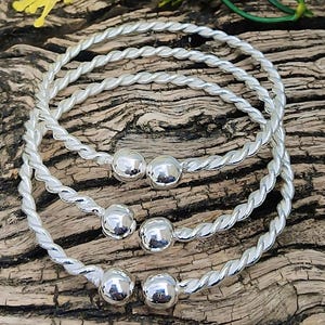 May include: Three silver-colored, twisted-rope style bangles with a smooth, spherical bead at each end. The bangles are displayed on a rustic, wooden surface, with a sprig of yellow flowers and green leaves in the background.