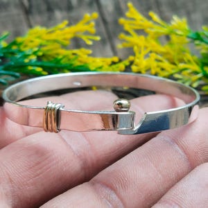 May include: Silver and gold-tone bangle bracelet. The bracelet features a hinged clasp with a small gold-tone ball and a section wrapped in gold-tone wire. The bracelet is displayed against a background of green and yellow foliage.
