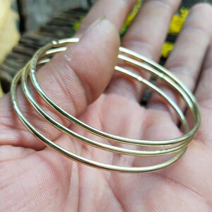 May include: A set of three thin, polished gold-tone metal bangles. Each bangle features a smooth, circular design with a reflective surface. The three individual rings are stacked together, creating a layered appearance with a consistent metallic finish.
