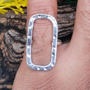 May include: A silver-colored ring with a rectangular, open-center design. The ring has a hammered texture and is worn on a finger. The background includes yellow and green floral accents.